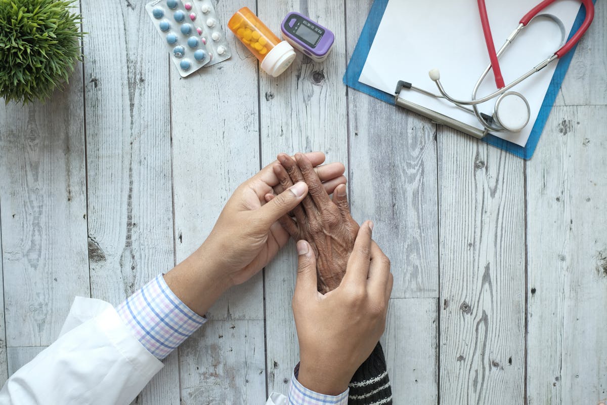 Avaliação Geriátrica Ampla: médica segura a mão do paciente; ao lado, medicamentos, oxímetro, prancheta e estetoscópio